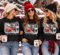 Three models wearing black Small Town Christmas graphic sweatshirts in a snowy holiday setting